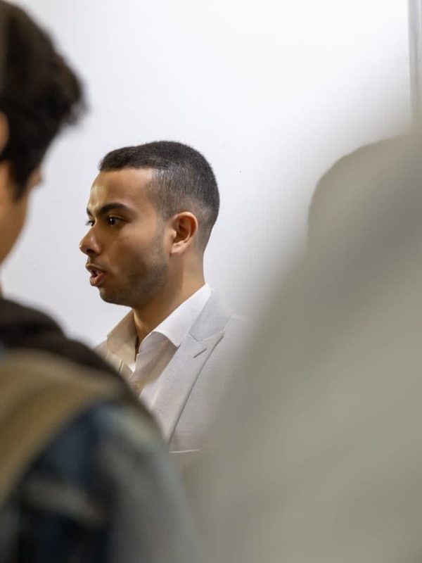 Ali Ahmed Samir speaking at an event, in a light jacket and white shirt, seen between two people in the foreground